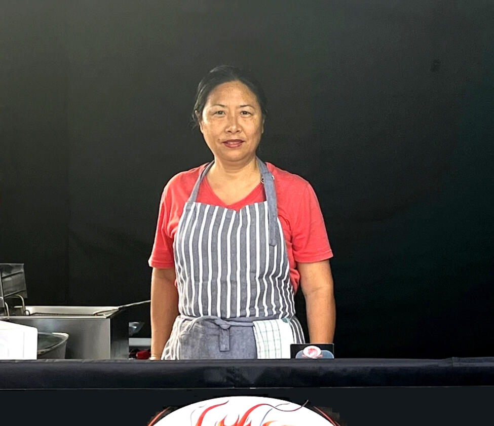 Portrait of Helen, the chef behind Oriental Savouries, standing at the market stall with a dark background and part of the dragon logo visible.